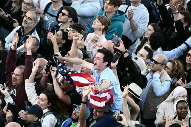 Multidão saúda o papa Leão 14 após o anúncio de sua eleição, com um homem segurando a bandeira dos Estados Unidos