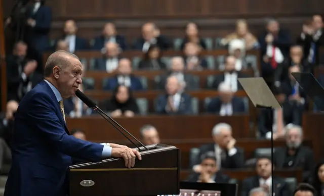 Erdoğan kürsüde konuşurken yandan görünüyor, geri planda flu AKP'liler koltuklarında amfi gibi bir alanda onu dinliyor.