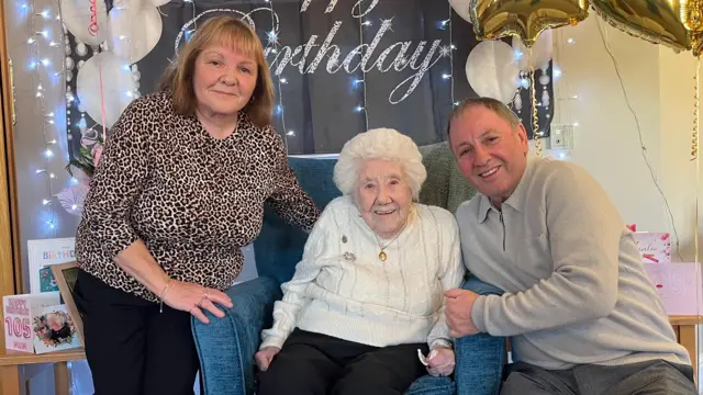 Sue Corr está a la izquierda. Tiene el pelo castaño hasta los hombros y lleva una blusa con estampado de leopardo y pantalones negros. En el centro está Elsie Wilkins, sentada en una silla, con el pelo blanco y un jersey blanco y pantalones negros. A la derecha está Brian Wilkins, con el pelo castaño corto y un jersey gris y pantalones.