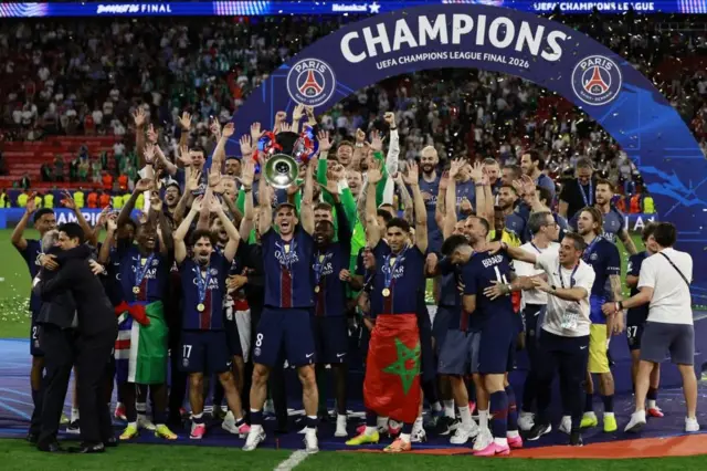 競技場のピッチで「CHAMPIONS」と書かれたアーチを前に、選手や関係者たちがトロフィーを掲げて喜んでいる