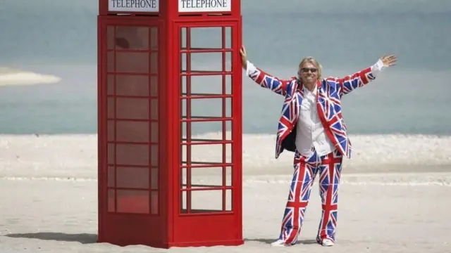 ਰਿਚਰਡ ਬ੍ਰਾਇਨ ਇੱਕ ਲਾਲ ਟੈਲੀਫੋਨ ਬੂਥ ਕੋਲ ਖੜ੍ਹੇ ਹਨ ਅਤੇ ਯੂਨੀਅਨ ਜੈਕ ਦੇ ਪ੍ਰਿੰਟ ਵਾਲਾ ਕੋਟ ਪੈਂਟ ਪਾਇਆ ਹੈ