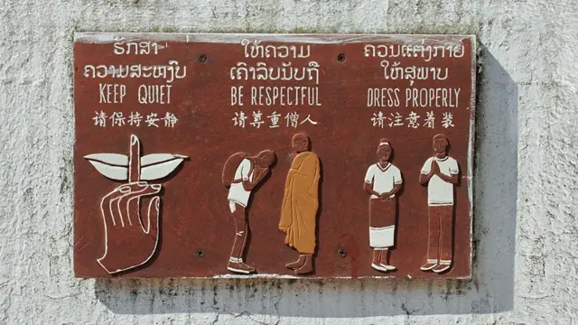 Placa na parede com orientações aos turistas sobre como devem se comportar, escritas em laociano, inglês e chinês, além de ilustrações