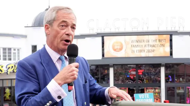 Nigel Farage está de pie frente al muelle de Clacton con un micrófono. Se puede ver un cartel de "Vote por la Reforma" al fondo y las atracciones están detrás de él. Lleva traje azul, corbata azul claro y camisa a cuadros rosa pálido y azul.