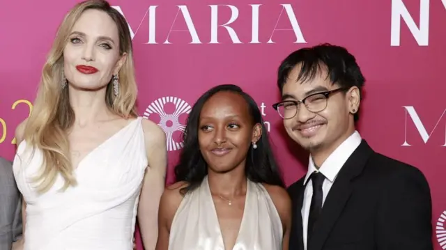 Angelina Jolie, Zahara Marley Jolie és Maddox Chivan Jolie-Pitt ünnepi öltözékben mosolyog a kamerába egy rózsaszín fal előtt.