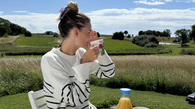 Vilma Larsson tomando uma xícara de chá ou café ao ar livre, em um dia ensolarado, com vista para o campo atrás dela