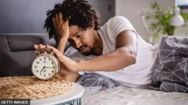 Aworan ọkunrin to fi ọwọ kan le aago orii tabili to si gbe ọwọ keji siwaju ori rẹ