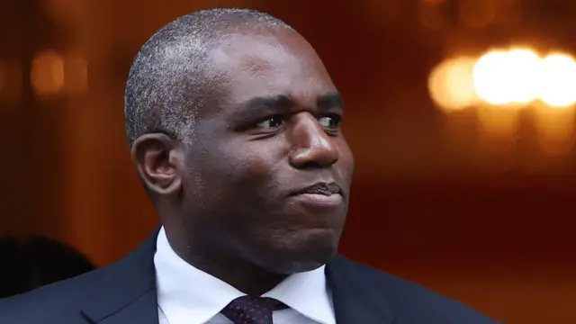 David Lammy tiene cabello gris muy corto y viste una camisa blanca, una chaqueta de traje azul marino y una corbata azul marino con manchas rojas.