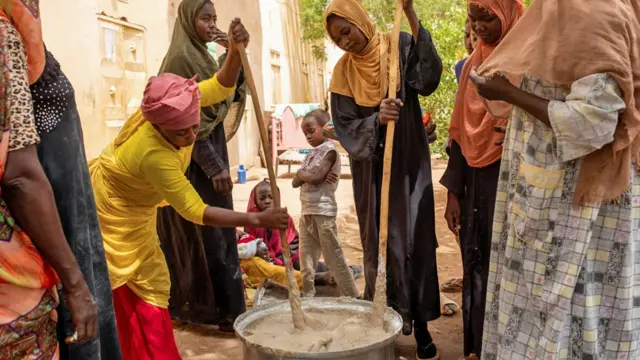Des femmes soudanaises d'une cuisine communautaire remuent du porridge dans un pot métallique géant avec de longs becs dans une cour à Omdurman, au Soudan - 2024.