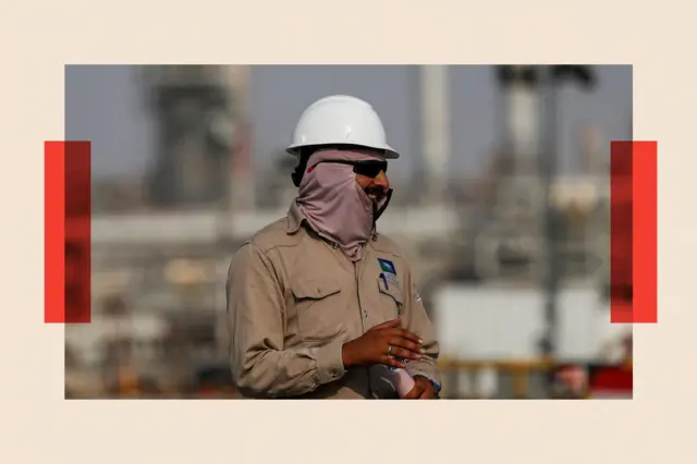 An oil rig worker wearing a white hard het and khaki boiler suit looks away from the camera
