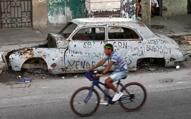 Un hombre maneja una bicicleta junto a un auto destartalado en una calle en Cuba.