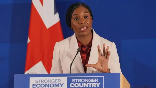 青い幕とイギリス国旗を背に、白いスーツ姿のベイドノック氏が演台を前に、手を開きながら話している。演台には「より強い経済、より強い国」と書かれている