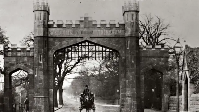 Fotografía de época en blanco y negro del Arco de Altrincham, procedente del archivo del Ayuntamiento de Trafford. Un hombre a caballo y en un carro pasa bajo el arco de ladrillo. Tiene dos arcos de ladrillo más pequeños a cada lado y una puerta metálica levantada.