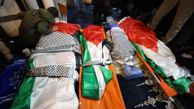 Uno de los niños sobrevivientes de la familia Bani Odeh se agacha al lado de cuatro cuerpos envueltos en banderas palestinas antes de su entierro en Tammun, Cisjordania ocupada (15 de marzo 2026)