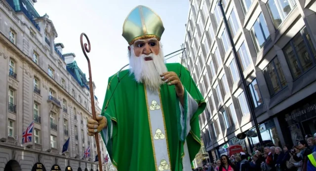 Kukła przedstawiająca św. Patryka, którą niosą uczestnicy marszu. W tle widać brytyjskie flagi i beżowe budynki.