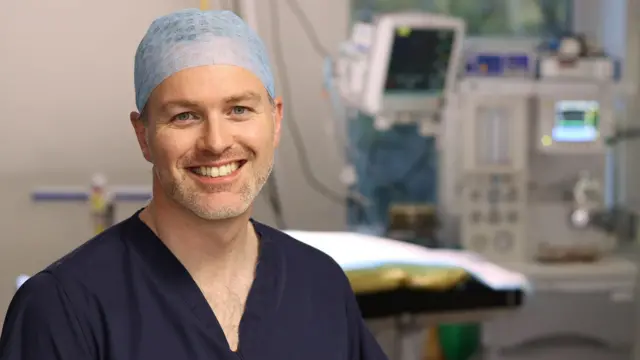 Um homem sorri em uma sala de cirurgia, usando uma touca de cabelo e um uniforme médico azul-marinho.