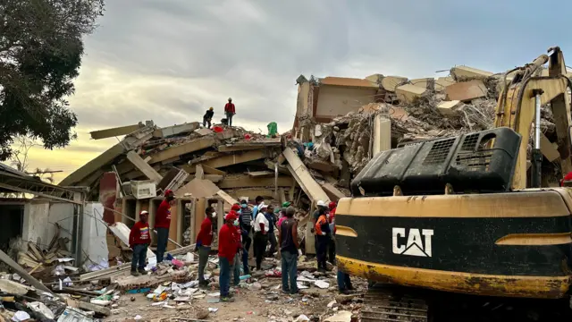 Rescatistas trabajando al atardecer para remover los escombros del conjunto residencial. Hay una grúa a un lado de ellos.