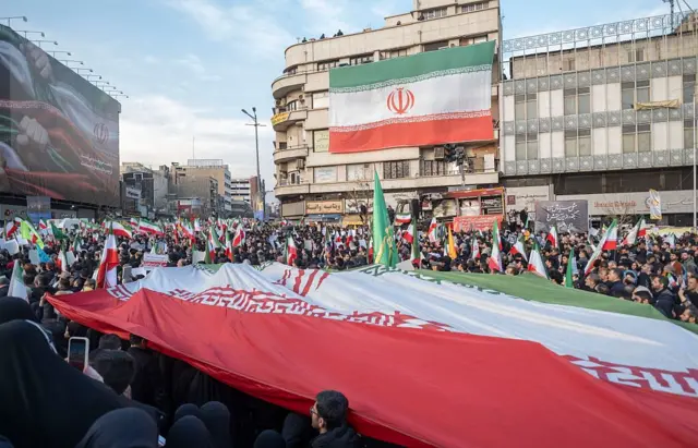 Большая толпа с иранскими флагами, на переднем плане люди держат большой флаг, также большой флаг висит на здании. На другом здании виден экран, на нем видны руки, держащие полоски ткани в цветах флага Ирана.