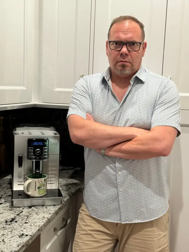 Andrew Buckley é um homem branco, com o cabelo claro e bem curto. Ele usa óculoscbet universitygrau, veste camisa clara e calça bege e aparececbet universitybraços cruzados ao ladocbet universityuma máquinacbet universitycafé