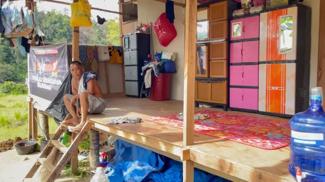 Seorang bapak lanjut usia, bertelanjang dada dan mengenakan celana pendek serta sandal jepit, bajunya diletakkan di pundak kirinya, duduk di teras huntara mandiri di Batu Busuk, Sumatra Barat, yang terbuat dari kayu, dibangun warga dari sumbangan para donatur.