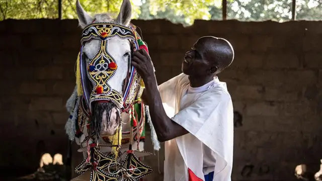 A man attends to a horse dressed in a colourful mask.