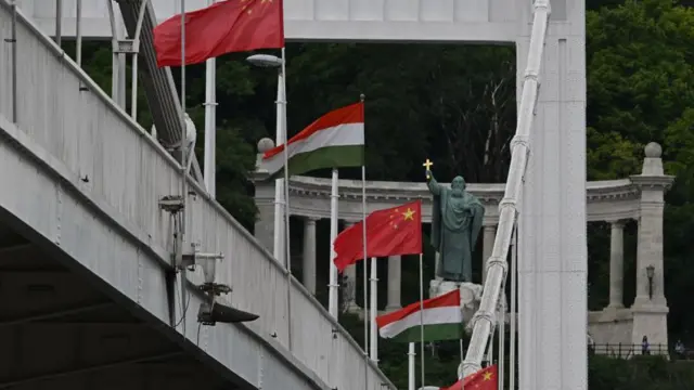 Kínai és magyar zászlók sorakoznak egy hídon Budapesten, a háttérben egy szobor és oszlopos építmény látható a Gellért-hegy alján.