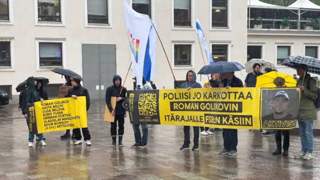 На фото видны около 10 человек, которые стоят под дождем и держат флаги, плакаты и баннер в поддержку Голикова и других соискателей убежища в Финляндии
