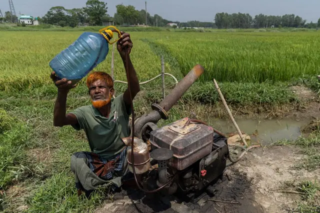 ফসলের ক্ষেতে সেচের পাম্পের সামনে বসে একজন ব্যক্তি একটি প্লাস্টিকের বোতল উঁচিয়ে দেখছেন
