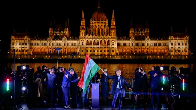 Péter Magyar, lider opozycyjnej partii Tisza, trzyma flagę narodową po ogłoszeniu częściowych wyników wyborów parlamentarnych w Budapeszcie podczas wiecu na świeżym powietrzu wzdłuż Dunaju, z podium i węgierską flagą przed oświetlonym budynkiem parlamentu.