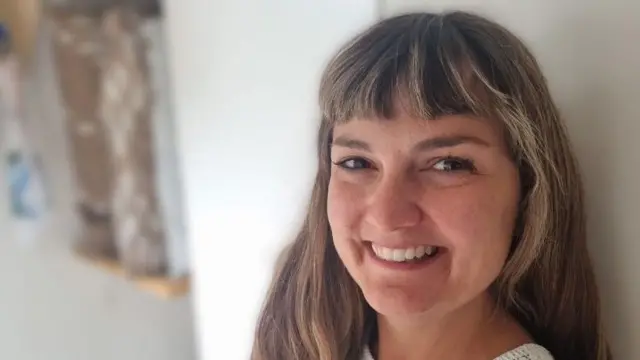 Une femme aux longs cheveux bruns clairs et ondulés, avec une frange, sourit à la caméra. Elle se tient debout devant un mur blanc, avec des étagères à sa gauche.