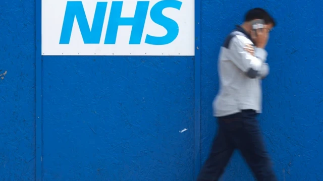یک مرد در حال صحبت با موبایل از مقابل دیوار آبی رنگ که روی آن نوشته NHS عبور می‌کند
