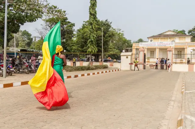 Une femme avec le drapeau béninois autour de sa taille, marche sur une route avec devant elle un bâtiment.