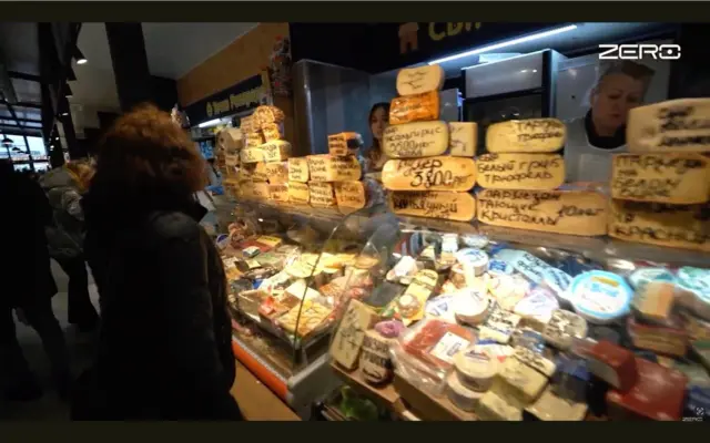 На ринку перед скляною вітриною з сирними головами стоїть покупчиня в темній куртці. На полицях – десятки сортів сиру з рукописними цінниками; за прилавком видно холодильні шафи. Усередині залу багато світла та людей.