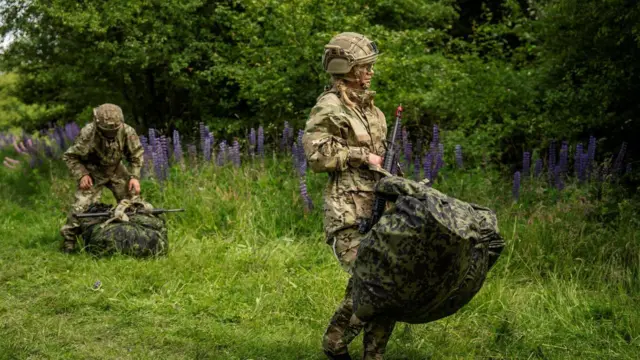 Kobieta służąca w duńskiej armii, ubrana w mundur wojskowy i hełm, niesie karabin i dużą torbę