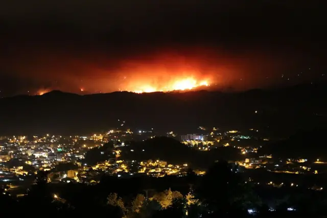gece vakti bir kent ve arkasındaki dağlardan yükselen kırmızı alevler