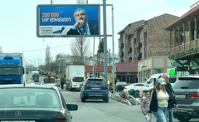 Автомобили на уходящей вдаль улице, которую переходит семья - муж, жена, ребенок. Над улицей на столбе выставлен баннер с портретом седого, но достаточно молодого мужчины, задумчиво глядящего вдаль, упираясь рукой в подбородок. 