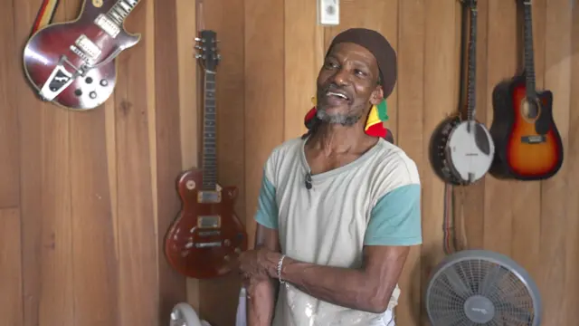 El músico de calipso Mateo Waite posa frente algunos de sus instrumentos de cuerda que cuelgan en una pared de madera dentro de su casa en Puerto Viejo de Talamanca.