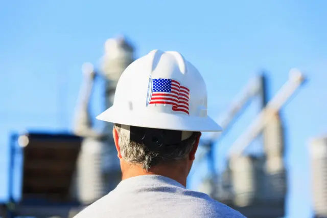 Homem com capaceteesportes da sorte piscou pagoutrabalho que tem bandeira dos EUA; ele observa planta industrial