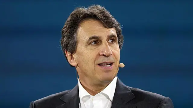 An image of businessman Marc Rowan looking to the right with a microphone attached to his face, while dressed in a black suit and sat in front of a blue backdrop. 