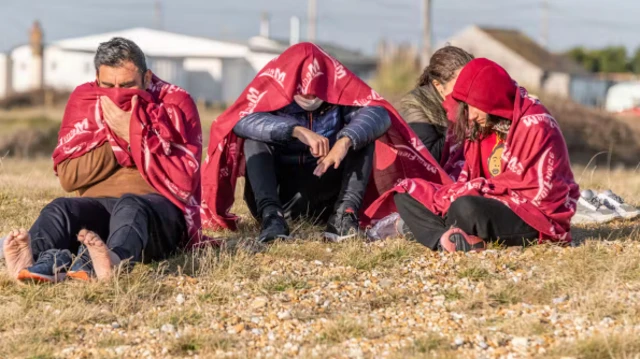  ثلاثة مهاجرين يفترشون الأرض ويبدو عليهم البرد إذ يضعون بطانيات حمراء اللون على أكتافهم.