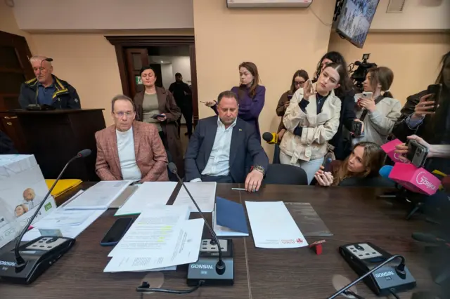 У залі засідань за столом із мікрофонами сидять Єрмак і Фомін перед паперами. Позаду й праворуч — група журналістів з камерами та телефонами, спрямованими на них. На столі видно документи, телефон і обладнання для запису звуку, у фоні — двері та люди біля входу.