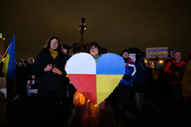 Вечірня акція підтримки: люди стоять на площі з прапорами і плакатами. У центрі – велике серце, поділене на кольори прапорів України та Польщі, як символ солідарності між країнами.
