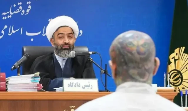 امیرحسین مقصودلو ایستاده در برابر قاضی دادگاه انقلاب اسلامی که پشت میز چولی نشسته است و لباس روحانی به سر دارد