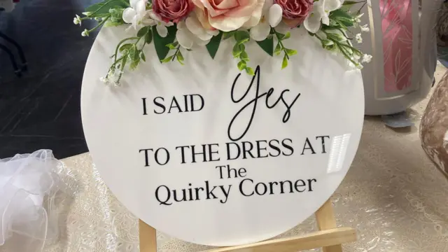 Un gran cartel circular blanco dice: «DIJE SÍ AL VESTIDO EN LA ESQUINA PECULIAR». Un ramo de flores lo cubre.