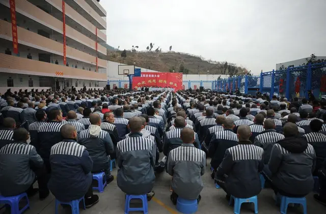 चीन के चुआंशी जेल में जेल की वर्दी पहने और प्लास्टिक के स्टूल पर बैठे कैदियों की कतारें