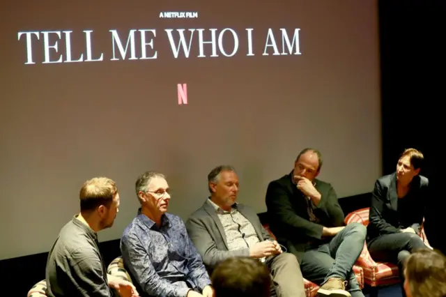Cinco hombres sentados en sillones de piel, uno al lado del otro. Todos van vestidos con camisa informal, pantalones vaqueros y zapatos casuales. Al fondo hay una pantalla grande y blanca donde se puede leer "Tell Me Who I Am", en español, "Dime quén soy" y debajo el logo de la plataforma Netflix, una letra N mayúscula en color rojo. 
