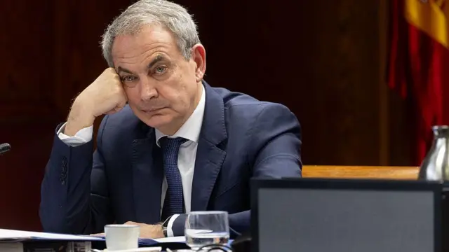 Zapatero con la cabeza apoyada en el puño y rostro cansado. Viste un traje azul marino y camisa blanca.
