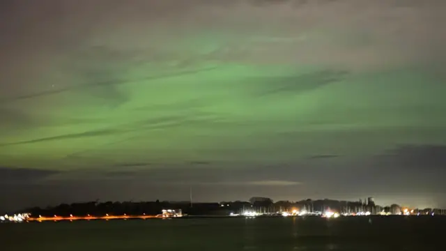 El mar de noche es negro y a lo lejos se ve el límite del pueblo costero. Luces centelleantes adornan el puerto y los barcos. El cielo se tiñe de verde y gris.