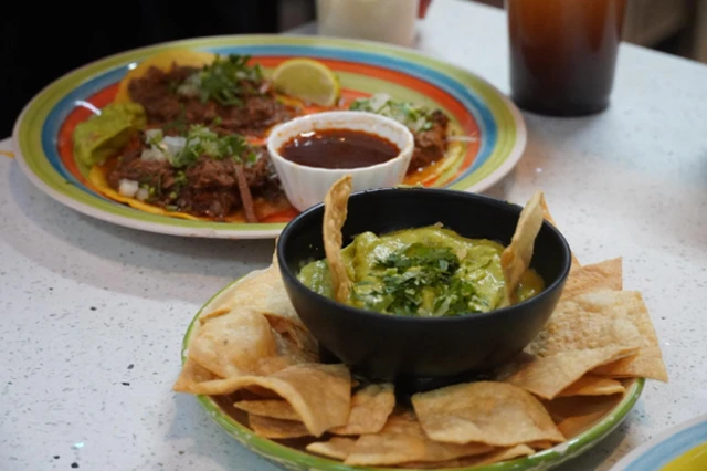 En primer plano se ve un plato de guacamole con tortillas y al fondo unos tacos de birria. 