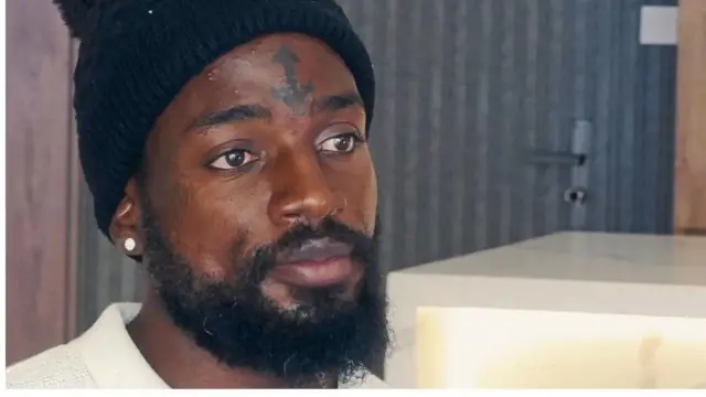 Troy to n ba Charles Mwesigwa ṣiṣe tẹlẹ ree, o de fila dudu, o fi ami kan siwaju ori rẹ. Troy fi yẹti seti osi rẹ, o ni irungbọn to pọ.