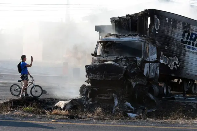 Un hombre en bicicleta toma una foto junto a un camión incendiado en Jalisco